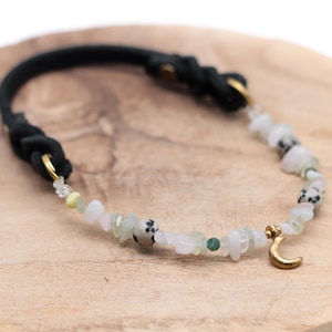 K&ouml;nnte beinhalten: Ein schwarzes Lederarmband mit einem goldenen Halbmond-Anh&auml;nger und wei&szlig;en, gr&uuml;nen und schwarzen Edelsteinperlen.