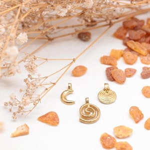 K&ouml;nnte beinhalten: Drei goldfarbene Anh&auml;nger mit verschiedenen Designs: eine Mondsichel, eine Spirale und ein Kreis mit einem Spiralmuster. Die Anh&auml;nger liegen auf einer wei&szlig;en Oberfl&auml;che mit kleinen Bernsteinst&uuml;cken, die um sie herum verstreut sind.