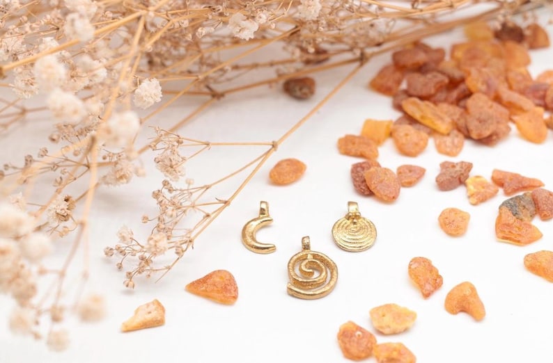K&ouml;nnte beinhalten: Drei goldfarbene Anh&auml;nger mit verschiedenen Designs: eine Mondsichel, eine Spirale und ein Kreis mit einem Spiralmuster. Die Anh&auml;nger liegen auf einer wei&szlig;en Oberfl&auml;che mit kleinen Bernsteinst&uuml;cken, die um sie herum verstreut sind.