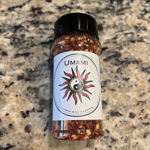 May include: A small glass jar with a black lid filled with red and brown flakes. The label reads "UMAMI" with a black and white yin yang symbol inside a sun with red rays. The label also reads "FARMER MICK'S GARDEN".