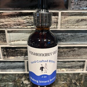 May include: A 2 ounce bottle of Fashionably Old Hand-Crafted Bitters. The bottle has a white label with blue text and a cartoon illustration of a couple in vintage clothing. The text on the label reads "Fashionably Old Hand-Crafted Bitters 2 oz Making bitters better."