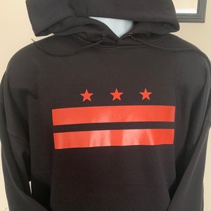 May include: Black hooded sweatshirt with a red and orange graphic design. The design features three stars above two horizontal stripes. The sweatshirt has a drawstring hood and long sleeves.
