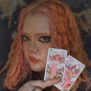 May include: Two rectangular tarot cards featuring intricate, colorful illustrations of figures in pink and red tones. The cards are held in a hand adorned with a metallic ring featuring a blue stone, set against a dark background with star patterns.
