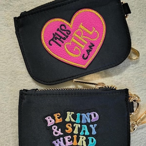 May include: Two black coin purses with gold zippers and key rings. One purse has a pink heart with the text "THIS GIRL CAN" in yellow. The other purse has the text "BE KIND & STAY WEIRD" in various colors.