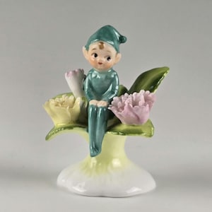 Könnte beinhalten: Eine Porzellanfigur eines kleinen Elfen mit blaugrünem Hut und Outfit, der auf einem blassgelben Sockel sitzt. Der Elf ist von modellierten Blumen in Blassgelb und zartem Rosa umgeben, mit grünen Blättern, die aus dem Sockel herausragen.