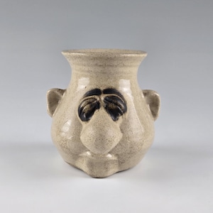 May include: A tan ceramic vase with a face design. The vase has a wide, round body with a flared rim and two small handles. The face features black eyes and a prominent nose. The vase is approximately 8 inches tall.