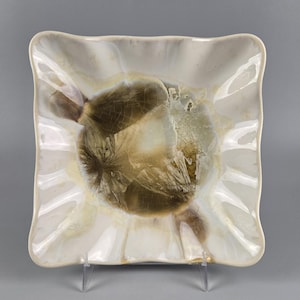 May include: Square ceramic dish with a ruffled edge. The dish features a cream-colored exterior with a central design of brown and cream crystalline formations. The dish is displayed on a clear stand against a gray background.
