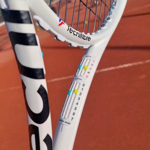 以下が含まれることがあります： フレームに「Tecnifibre」というブランド名が印刷された白黒のテニスラケット。ラケットには、ハンドルに青、黄、赤のインジケーターが付いたスコアキーピングデバイスが付いています。