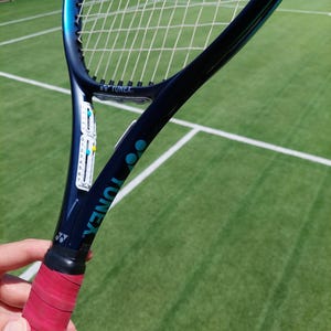 以下が含まれることがあります： 赤いグリップが付いた青と黒のヨネックスのテニスラケット。ラケットは緑のテニスコートにあります。