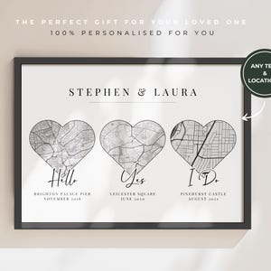 May include: A black framed print with three heart-shaped map designs. The print includes the names "Stephen & Laura" above the hearts, with the words "Hello", "Yes", and "I Do" below each map. The text "The perfect gift for your loved one" is at the top.