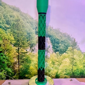 Puede incluir: Objeto alto, verde y negro, impreso en 3D, con forma cilíndrica. La sección superior es verde sólido, mientras que la sección media presenta un diseño de celosía de diamantes en verde y negro. Se apoya sobre una base circular con superficie reflectante.