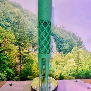 Puede incluir: Un objeto decorativo verde y alto con una parte superior cilíndrica con un patrón de diamantes. La sección media tiene un diseño de celosía y se apoya en tres patas delgadas. El objeto se exhibe sobre una base redonda y espejada.