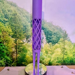 Puede incluir: Un objeto decorativo alto y morado con una parte superior cilíndrica y un diseño de celosía de diamantes en el cuerpo. Se apoya sobre dos patas delgadas y una base redonda negra. El fondo presenta un paisaje con árboles y un cielo morado claro.