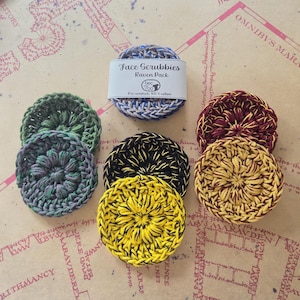 May include: A collection of handmade face scrubbies in various colors, including green, blue, yellow, and burgundy. The scrubbies are round and crocheted, with a label that reads "Face Scrubbies Raven Pack". They are made of pre-washed 100% cotton.