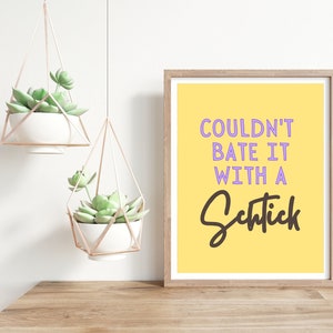 May include: A yellow background with the text "COULDN'T BATE IT WITH A SCHTICK" in purple and black lettering. The text is framed in a light brown wood frame.