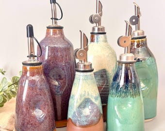 May include: Five ceramic oil dispensers with metallic pour spouts and black caps. The bottles feature varied reactive glazes in shades of deep purple, cream, teal, and brown, with smooth, rounded bodies and distinct color transitions on each piece.