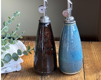 May include: Two ceramic oil dispensers with tapered bodies and stainless steel pour spouts. One dispenser features a glossy dark brown glaze with subtle speckles, while the other has a textured blue glaze with lighter blue and grey variations.