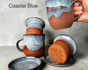 May include: Ceramic tea mug set in Coastal Blue featuring a terracotta base with a blue glazed top. The set includes a mug with a handle, a removable tea infuser with small drainage holes, and a matching saucer. The text Coastal Blue is visible.
