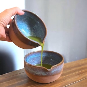 May include: A ceramic matcha bowl with a brown exterior and blue interior is being used to pour green matcha tea into another bowl. The bowls are on a wooden surface. The tea is a vibrant green color.
