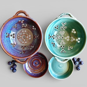 May include: Two ceramic colanders, one blue-purple and one teal-green, with matching small bowls. The colanders have decorative handles and patterned holes. Blueberries are scattered around the bowls, suggesting their use for washing and serving fruit.