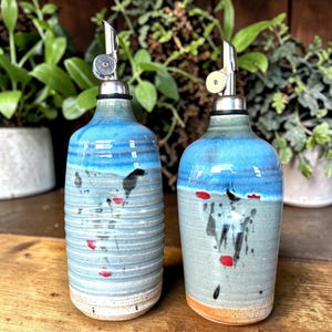 May include: Two ceramic oil dispensers with a blue and grey glaze, featuring a ribbed texture on the left bottle and a smooth finish on the right. Both bottles have a silver-colored metal pour spout and are decorated with small red and black accents.