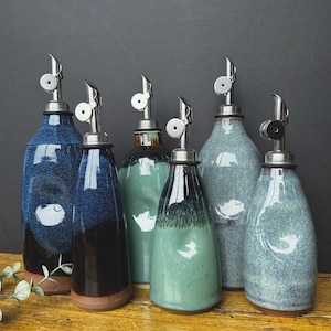 May include: A collection of ceramic oil bottles with stainless steel spouts. The bottles are in various shades of blue, green, and gray, with a speckled glaze finish. Each bottle has a unique shape and design, with a brown base.