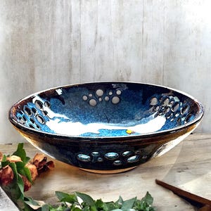 May include: A handcrafted ceramic bowl with a unique design. The bowl features a dark blue exterior with a lighter blue and white interior. It has a series of circular cutouts around the rim, adding a decorative element. The bowl is suitable for serving or display.