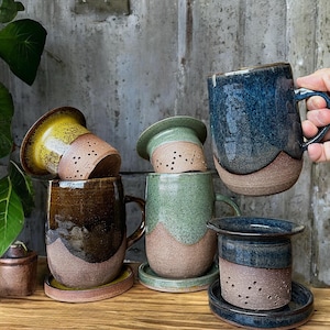 May include: A collection of handmade ceramic mugs and tea infusers in various colors, including brown, green, and blue. Each mug has a unique glaze and design, with matching saucers. The tea infusers have small holes.