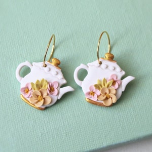 Può includere: Un paio di orecchini a forma di teiera bianca con dettagli dorati e fiori rosa e gialli. Gli orecchini sono appesi ad anelli dorati.