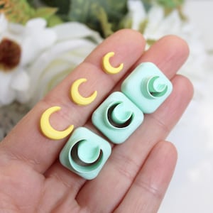 Può includere: Una mano tiene diversi tagliapasta in argilla polimerica a forma di mezzelune. Ci sono tre mezzelune gialle e tre tagliapasta quadrati con un disegno a mezzaluna. I tagliapasta sono verde chiaro.