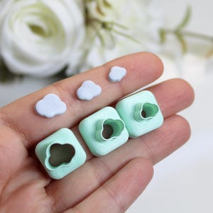 Op de afbeelding: Een hand houdt drie vierkante, lichtgroene koekjessnijders met wolkvormige uitsparingen en drie kleine, lichtblauwe, wolkvormige decoraties vast. Witte bloemen en groen zijn op de achtergrond.