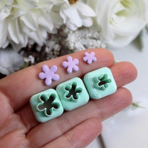 Könnte beinhalten: Eine Hand hält drei quadratische, hellgrüne Ausstecher in Blumenform und drei kleine, hellviolette Blumendekorationen. Weiße Blumen sind im Hintergrund.