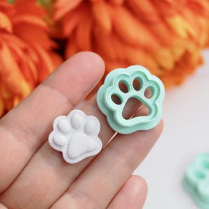 Op de afbeelding: Twee pootvormige koekjessnijders, één witte en één lichtgroene, worden getoond. De witte pootafdruk is een afgewerkt product, terwijl de groene een snijder is. Oranje bloemen staan op de achtergrond.