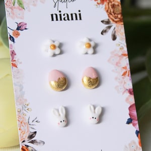 Könnte beinhalten: Eine Karte mit sechs Paar Ohrsteckern mit Ostermotiven. Die Ohrringe umfassen weiße Blumen, rosa und goldene Eier und weiße Hasenmotive. Die Karte trägt den Text "studio niani" und florale Akzente.