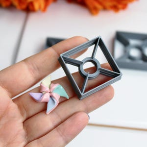 Könnte beinhalten: Ein grauer, quadratischer Ausstecher in Form eines Windrads mit einem zentralen Kreis und einem kleinen, bunten Windrad. Der Ausstecher wird in einer Hand gehalten, ein zweiter Ausstecher ist im Hintergrund zu sehen. Orangefarbene Blumen sind im Hintergrund.