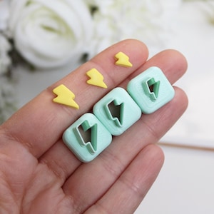 Può includere: Una mano tiene diversi piccoli oggetti gialli a forma di fulmine e tre taglierini quadrati verde chiaro con ritagli a forma di fulmine. Gli oggetti sono probabilmente per l'artigianato o la cottura.