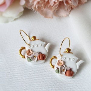 Puede incluir: Un par de pendientes de aro dorados con dijes de tetera blanca. Las teteras tienen pequeños detalles florales y de calabaza en tonos naranja, rosa y verde.