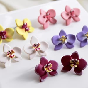 Könnte beinhalten: Handgefertigte Ohrstecker in Form von Orchideenblüten in verschiedenen Farben: Gelb, Rosa, Lila, Weiß und Burgunderrot. Die Ohrringe sind auf einer weißen quadratischen Fliese angeordnet und zeigen ihr detailliertes Blumendesign.