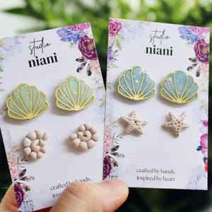 Könnte beinhalten: Zwei Karten mit Ohrringen werden in einer Hand gehalten. Jede Karte zeigt ein Muschel- und Seestern-Design. Die Muschelohrringe haben eine goldene Umrandung mit einem hellgrünen oder blauen Zentrum. Die Seestern- und Spiralmuschel-Ohrringe sind hellbeige. Die Karten haben florale Akzente und den Text "studio niani".