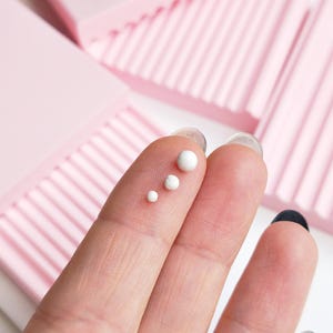 Könnte beinhalten: Drei kleine, kugelförmige weiße Perlen unterschiedlicher Größe auf einem Finger, platziert vor mehreren rechteckigen, hellrosa Blöcken, die eine Reihe paralleler, vertikaler Rillen auf ihren Oberflächen aufweisen.