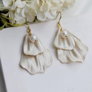 Könnte beinhalten: Cremefarbene, geschichtete Blütenblatt-Ohrringe mit einem kleinen Perlenakzent und goldfarbenen Haken. Die Ohrringe werden auf einer weißen Oberfläche mit weißen Blumen im Hintergrund präsentiert. Die Ohrringe sind etwa 5 cm lang.