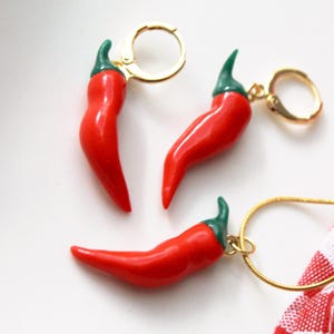 Könnte beinhalten: Ein Schmuckset mit Chili-Pfeffer-Designs. Das Set enthält Ohrringe mit goldfarbenen Reifen und eine Halskette mit einer goldfarbenen Kette. Die Paprika sind rot mit grünen Stielen. Ein rot-weiß kariertes Tuch befindet sich im Hintergrund.