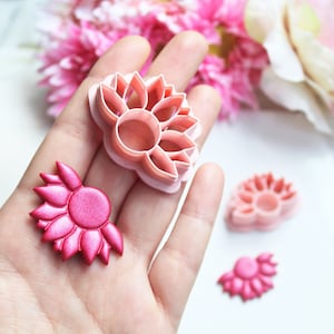 Peut inclure: Un ensemble de trois emportes-pièces en plastique de fleurs roses et blanches. Les emportes-pièces sont en forme de fleur de lotus. Les emportes-pièces sont montrés sur une main, avec une fleur de lotus en pâte à sucre rose à côté d'eux.