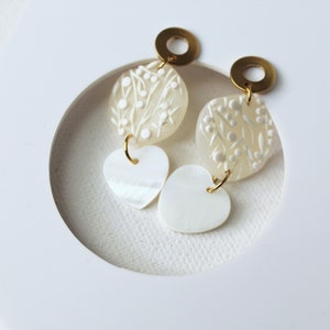 Könnte beinhalten: Ein Paar weiße Muschelohrringe mit floralem Design und einem herzförmigen Anhänger. Die Ohrringe haben goldfarbene Metallakzente.