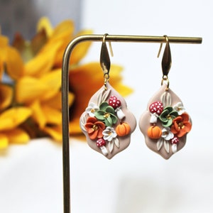 Op de afbeelding: Een paar hangende oorbellen met een lichtbruine, vierlobbige basis. Elke oorbel heeft een cluster van polymer clay bloemen in tinten oranje, wit en groen, samen met kleine, rood-witte paddenstoelen en een miniatuur oranje pompoen.