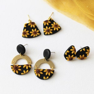 Könnte beinhalten: Drei Paar schwarze und goldene Ohrringe mit Sonnenblumen-Designs. Die Ohrringe sind aus Polymer Clay gefertigt und haben goldfarbene Metall-Verschlüsse.