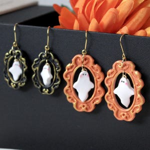 Op de afbeelding: Vier paar Halloween-oorbellen. Twee paar hebben een zwart-gouden frame en twee hebben een oranje frame. Elke oorbel heeft een witte spookbedel met roze wangen en goudkleurige oorhaken.
