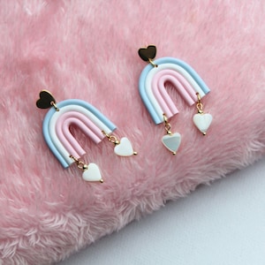 Könnte beinhalten: Ein Paar Regenbogenohrringe mit einem herzförmigen Anhänger oben und drei baumelnden herzförmigen Anhängern. Die Ohrringe sind aus Polymer Clay gefertigt und haben ein pastellfarbenes Rosa-, Weiß- und Hellblauschema.