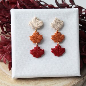 Puede incluir: Un par de pendientes colgantes con tres hojas de arce superpuestas en tonos crema, naranja y rojo.