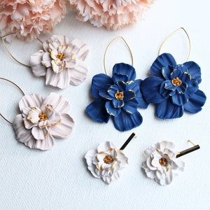 Könnte beinhalten: Drei Paar Blumenohrringe mit geformten Blütenblättern, deren Ränder und Mitten goldfarben bemalt sind. Ein Paar ist hellrosa mit goldenen Drahtreifen, eines ist dunkelblau mit goldenen Drahtreifen und eines ist weiß mit geraden goldenen Stiften.